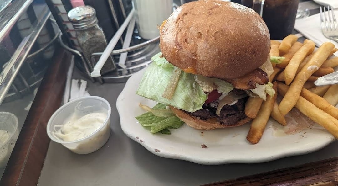 A delicious burger with lettuce, tomato, and a toasted bun, served with a side of crispy French fries and a small cup of mayonnaise at Crosstown Diner in Bronx, NY